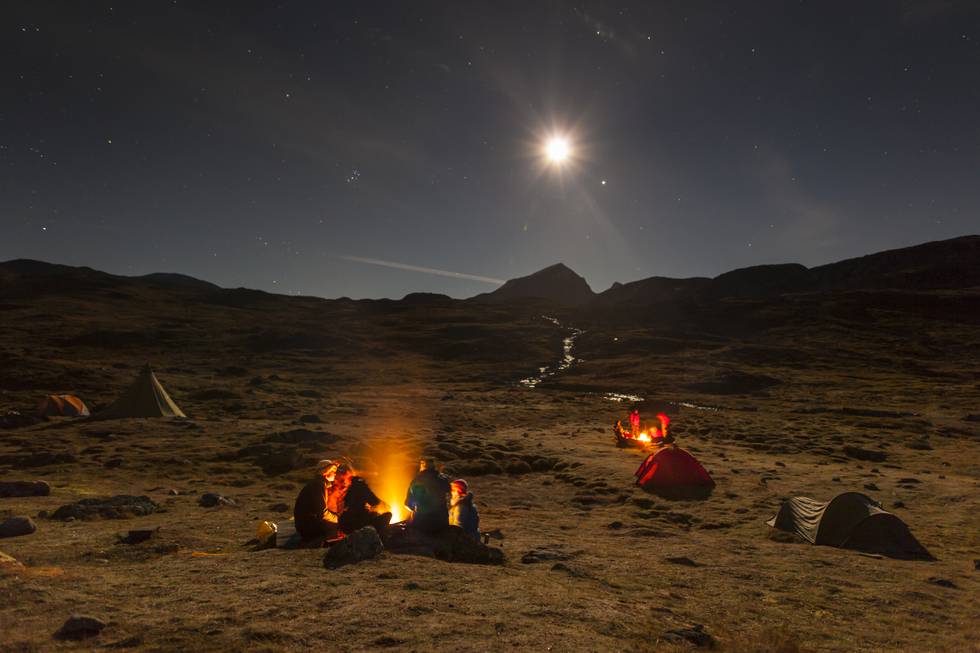 Mennesker rundt bål en kald høstkveld i Jotunheimen. Månen lyser opp og i bakgrunnen ses Store Smørstabbtind.
 Foto: © Espen Haagensen / NTB